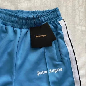 Blå Palm Angels shorts med vit rand - Snygga blå shorts från Palm Angels med vit och svart rand längs sidorna. Elastisk midja och broderad logga framtill. Perfekta för en chill och sportig look.