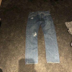Levi's 501 blå jeans W32 L32 - Klassiska Levi's 501 jeans i blå denim med rak passform och normal midja. Snygga slitningar och hål på ena benet ger en cool, avslappnad vibe. Fem fickor och knappgylf. Perfekt för dig som gillar vintage och streetstyle.