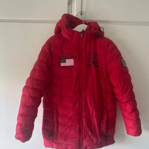 Röd pufferjacka från Vinson Polo Club - Röd pufferjacka från Vinson Polo Club med huva och dragkedja. Jackan har broderade detaljer på bröstet, inklusive amerikansk flagga och logga, samt patch på ärmen. Perfekt för kyliga dagar och har justerbar huva.