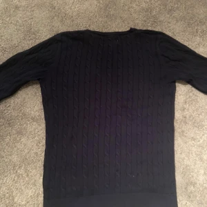 Mörkblå stickad tröja Hampton Republic - Snygg mörkblå stickad tröja från Hampton Republic med klassiskt kabelstickat mönster och rund hals. Tröjan har ett broderat vitt emblem med siffran 27 på bröstet och ribbade muddar vid ärmslut och nederkant. Perfekt för dig som gillar preppy stil.