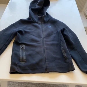 Svart hoodie från Nike med dragkedja - Svart hoodie från Nike i storlek L, med hel dragkedja framtill och två fickor med dragkedja. Tröjan har huva och är tillverkad i mjukt fleecematerial, perfekt för chill eller träning. Nike-logga på bröstet ger en clean och sportig vibe. Är lite solbränd och någon fläck.