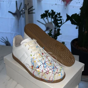 Maison Margiela sneakers med färgstänk - Unika vita sneakers från Maison Margiela med coola färgstänk i rött, blått, gult, grönt och svart. Skorna har klassisk snörning, rund tå och är tillverkade i skinn. Perfekt för dig som vill sticka ut med en konstnärlig vibe.