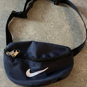 Mörkblå magväska från Nike - Snygg mörkblå magväska från Nike med klassisk vit logga framtill. Väskan har justerbart svart band och dragkedja med dubbla zippar. Perfekt för att bära det viktigaste och enkel att slänga över axeln.
