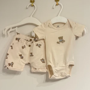 Beige shorts och body med nallemönster H&M baby - Supersöta beige babyshorts från H&M med gulligt nallemönster i brunt och blått. Shortsen har elastisk midja och små knappar framtill. Tillverkade i mjuk ekologisk bomull, perfekta för de allra minsta.