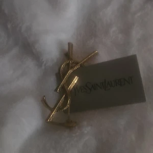  guldigt brosch - Säljer en ikonisk brosch i guldig metall med loggan. Snyggt texturerad yta som ger extra lyxig känsla. Perfekt accessoar för att lyfta vilken outfit som helst med en touch av designer-vibe.