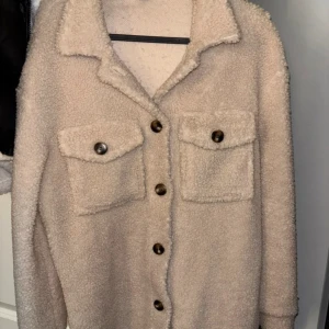 Beige teddyjacka med knappar - Säljer en beige teddyjacka med stora bruna knappar och två rejäla bröstfickor. Jackan har en klassisk krage och är långärmad, perfekt för dig som gillar mysiga och trendiga plagg. Materialet är fluffigt och ger en chill vibe. Jackan är använd ett fåtal gånger och är i storlek XS men är stor i storleken. 