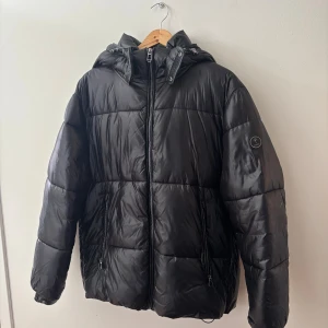 Svart pufferjacka från JOOP! med huva - Svart pufferjacka från JOOP! med stor logga på insidan, avtagbar huva och två dragkedjeförsedda fickor. Jackan är quiltad och tillverkad i återvunnen polyester, perfekt för kalla dagar. Snygg och stilren look med diskret JOOP!-märke på ärmen. 4500 ny pris säljer för 900kr Inga skavanker eller märken på den. Den är som ny för inte använt alls mycket då den blev lite stor för mig 