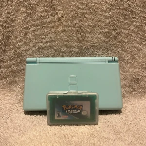 Nintendo DS Lite ingår fake Pokémon Emerald - Nintendo DS Lite i ljusblå färg, komplett med stylus ingår en fake Pokémon Emerald spelkassett. Konsolen och spelet är i gott skick, med minimalt slitage. Skärmen och knappar fungerar utmärkt, och spelet startar utan problem. Perfekt för retrospelare och samlare!