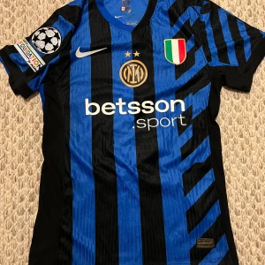 Inter Milan Calhanoglu #20 matchtröja - Inter Milan fotbollströja med Calhanoglu #20, tillverkad av Nike i lätt och ventilerande material. Klassisk blå och svart randig design med klubbmärke, sponsorer och Champions League-märke på ärmen. Kortärmad modell med snygga detaljer och guldinslag på sidorna.