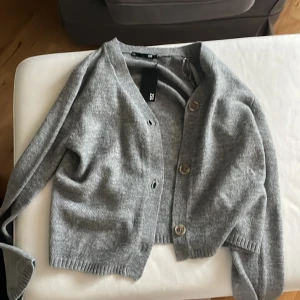 Grå kofta från 157 med knappar - Mysig grå kofta från 157 i storlek XS. Koftan har en klassisk v-ringad design med tre stora knappar framtill och ribbade muddar vid ärmslut och nederkant. Perfekt att slänga på sig när det är lite kyligt ute.