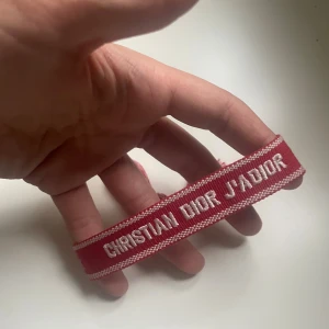 Röd armband Christian Dior J'Adior - Snyggt röd armband från Christian Dior med vit broderad text 'J'Adior'. Armbandet har två tofsar i ändarna och är gjort i ett mjukt tygmaterial. Perfekt accessoar för att lyfta din outfit med en lyxig touch.
