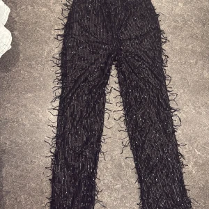 Svarta paljettbyxor från Gina Tricot - Unika svarta byxor från Gina Tricot med glittriga paljetter och fransdetaljer över hela ytan. Byxorna har rak passform och är perfekta för dig som vill sticka ut med en cool och festlig look. Materialet är syntet och de har en bekväm midja.