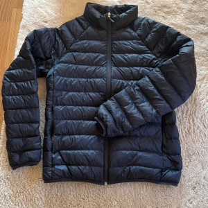 Svart pufferjacka med dragkedja - Säljer en svart pufferjacka med horisontella quiltade sektioner och hög krage. Jackan har hel dragkedja framtill och långa ärmar, perfekt för kyliga dagar. Enkel och stilren design som funkar till många olika outfits. Bra skicka nypris 799kr