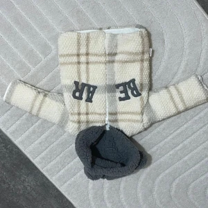 Beige teddyjacka med huva - Säljer en mysig teddyjacka i beige med rutigt mönster och mörkgrå huva. Jackan har dragkedja framtill och coola bokstäver på bröstet. Perfekt för dig som gillar chill och varm stil.