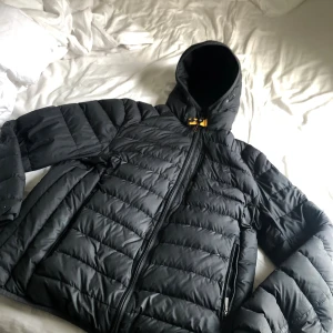 Svart dunjacka Parajumpers XL - Svart dunjacka från Parajumpers i storlek XL men passar L. Jackan är superlätt, har huva, gul detalj vid halsen och dragkedja framtill samt vid fickorna. Quiltad design och sportig look, perfekt för kalla dagar. Insidan är grå och har flera coola etiketter.