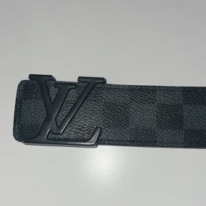 Svart rutigt bälte - Säljer ett stilrent svart bälte  med klassiskt rutigt mönster och en stor -logga i metall som spänne. Bältet har en slät och smal form, perfekt för att lyfta din outfit med en lyxig touch. Materialet är skinn med präglad struktur.