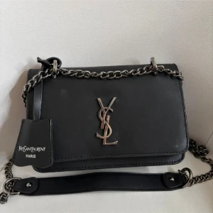 Svart axelväska - Snygg svart axelväska med klassisk YSL-logga i metall framtill. Väskan har en kedjerem med läderdetalj och flera fack inuti med vinrött foder. Stilren och lyxig design i slätt skinn, perfekt för dig som gillar exklusiva accessoarer.