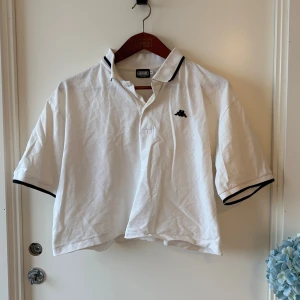 Vit pikétröja från Kappa x Episode - Vit pikétröja med svart Kappa-logga broderad på bröstet och svarta detaljer på krage och ärmslut. Tröjan har klassisk krage, korta ärmar och är tillverkad i mjuk bomull. Finns ett litet hål som syns knappt på andra bilden 