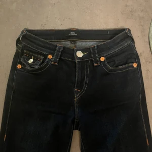True Religion mörkblå bootcut jeans Becca  - Snygga mörkblå jeans från True Religion med klassiska kontrastsömmar och ikoniska ficklock med silverfärgade knappar bak. Modellen har bootcut-snitt och låg midja, tillverkade i stretchigt jeansmaterial för skön passform. Perfekt för dig som gillar statement-jeans.