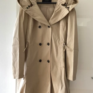 Beige trenchcoat från Esprit - Snygg beige trenchcoat från Esprit med dubbelknäppning, huva och knyt i midjan. Jackan har långa ärmar, svarta knappar och en klassisk, rak passform. 