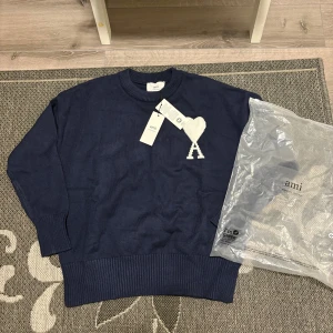 Mörkblå stickad tröja från Ami - Snygg mörkblå stickad tröja från Ami med rund halsringning och ribbade muddar. På bröstet finns ett vitt hjärta med bokstaven A. Perfekt för dig som gillar stilrena och trendiga plagg med en twist.