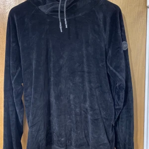 Svart Colmar hoodie i sammet - Svart hoodie från Colmar Originals i mjuk sammet med huva och snörning. Tröjan har lång ärm och ribbad nederkant med snygg asymmetrisk detalj baktill. Colmar-logga på ärmen ger en cool touch. Perfekt för dig som gillar stilren och bekväm streetwear.