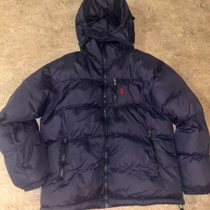 Mörkblå pufferjacka från Polo Ralph Lauren - Säljer en mörkblå pufferjacka från Polo Ralph Lauren med huva och dragkedjor framtill. Jackan har det klassiska röda Polo-logot broderat på bröstet, två sidofickor med dragkedja och är tillverkad i syntetmaterial. Perfekt för kalla dagar och har en schysst oversized vibe. passar M/S