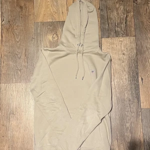 Beige hoodie från GANT med huva - Snygg beige hoodie från GANT i storlek S. Tröjan har en klassisk huva med snörning och GANT-logga broderad på bröstet. Tillverkad i mjuk bomull, perfekt för chill dagar. Långärmad och med ribbade muddar för extra komfort.