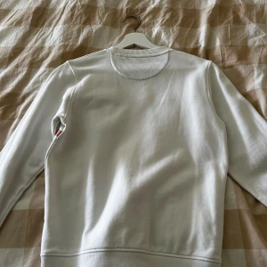 Vit sweatshirt från GANT med rosa detaljer - Snygg vit sweatshirt från GANT med rosa ränder och stor svart logga framtill. Tröjan har rund hals, ribbade muddar och är långärmad. Perfekt för dig som gillar klassisk stil med en modern touch.