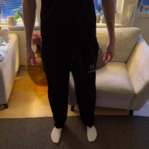 Svarta mjukisbyxor från Haglöfs - Storlek är XL men passar L. Svarta mjukisbyxor från Haglöfs med logga på vänster ben. Byxorna har elastisk midja med snörning och raka ben. Perfekta för chill eller träning, tillverkade i mjukt syntetmaterial som känns skönt mot huden.