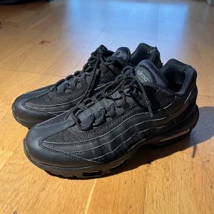 Nike Air Max 95 helsvarta - Nike Air Max 95 sneakers i helsvart färg med chunky siluett och synliga Air-enheter i sulan. Ovandelen är en mix av mesh och skinn med snygga vågformade paneler och snörning. Perfekt för dig som gillar streetwear och vill ha en clean look.
