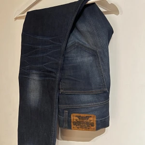 Crokers 308 mörkblå jeans - Säljer ett par croker 308 taperd jeans i mörkblå tvätt med snygga slitningar och whiskers på låren. Klassisk femficksmodell med brunt läderpatch bak och raka ben. Jeansen har kontrastsömmar och är tillverkade i slitstarkt denimtyg.
