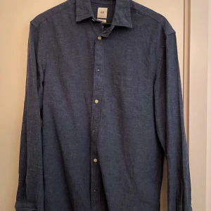 Mörkblå linneskjorta från H&M - Snygg mörkblå skjorta från H&M i linne med klassisk krage och knäppning framtill. Skjortan har långa ärmar och en bröstficka, perfekt för en chill och stilren look. Passar dig som gillar enkel och tidlös stil.