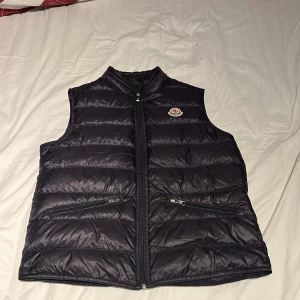 Svart dunväst från Moncler - Snygg svart väst  från Moncler med quiltad design och klassisk logga på bröstet. Västen har två fickor med dragkedja och hög krage. Perfekt att slänga över en hoodie för extra värme. Materialet är glansigt och känns lyxigt.