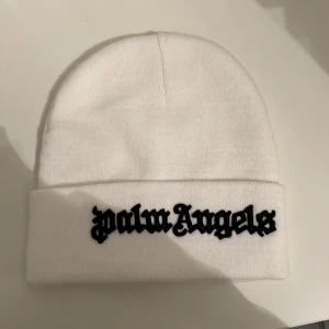 Vit mössa från Palm Angels - Säljer en clean vit mössa från Palm Angels med svart broderad logga framtill. Klassisk ribbstickad design med uppvikt kant. Perfekt accessoar för att lyfta din streetwear-look och hålla dig varm under kyliga dagar.