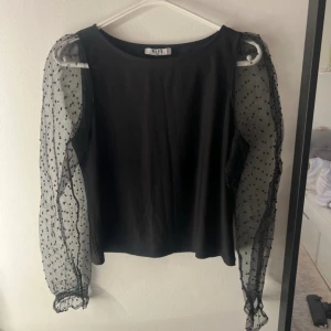 Svart blus med prickiga meshärmar från NA-KD - Säljer en svart blus från NA-KD med transparenta meshärmar som har små svarta prickar. Blusen har rund hals och lång puffärm med volangdetalj vid ärmslut. Kroppen är i mjuk bomull och ärmarna i tunn mesh. Perfekt för dig som gillar unika detaljer!