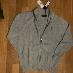 Grå stickad kofta - Säljer denna snygga gråa full zip koftan med röd logga.