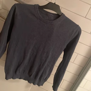 Mörkblå stickad tröja med rund hals - En enkel och stilren mörkblå stickad tröja med rundad hals och ribbade muddar. Tröjan har lång ärm och är gjord i ett mjukt stickat material som känns skönt mot huden. Perfekt för dig som gillar klassiska och cleana plagg.  har krympt något i tvätten och passar någon runt ca 146-152 cm.