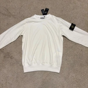 Vit Stone Island sweatshirt - Vit sweatshirt från Stone Island med klassisk rund hals och ribbade muddar. Tröjan har den ikoniska svarta och gula logotypen på vänster ärm. Tillverkad i mjuk bomull för skön komfort och clean look. Perfekt för dig som gillar stilrena plagg med street-vibe.