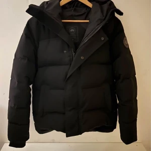 Canada Goose black label - Säljer en svart pufferjacka från Canada Goose med klassisk patch på ärmen. Jackan har huva, dragkedja och tryckknappar framtill samt stora fickor. Perfekt för kalla vinterdagar och riktigt snygg streetstil. ihopsydd under armen men inget som syns