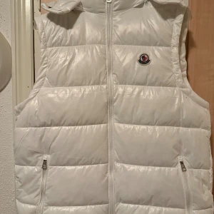 Vit dunväst från Moncler - Snygg vit dunväst från Moncler med klassisk logga på bröstet. Västen har quiltad design, dragkedja framtill och två fickor med dragkedja. Perfekt för lager-på-lager och ger en clean look. Insidan har coolt Moncler-tryck. aldrig använd