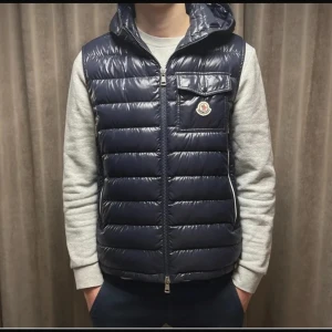 Mörkblå dunväst från Moncler - Snygg mörkblå dunväst från Moncler med huva och quiltad design. Västen har en bröstficka med lock och Moncler-logga, samt dragkedja framtill. Perfekt för lager-på-lager och streetstyle. Materialet är glansigt och känns lyxigt.