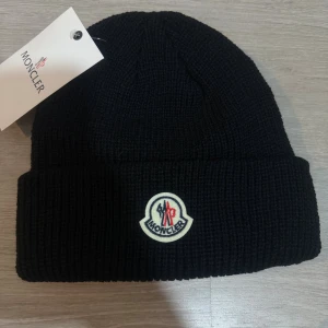 Moncler mössa  - Svart ribbstickad mössa från Moncler med klassisk uppvikt kant och broderad Moncler-logga framtill. Snygg och enkel design som passar till allt, tillverkad i mjukt stickat material för extra komfort.
