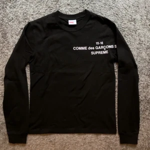 Supreme x Comme des Garçons svart tröja - Svart långärmad tröja från Supreme x Comme des Garçons med vit tryckt text på bröstet. Klassisk rund hals och ribbade muddar vid ärmslut. Perfekt för dig som gillar streetwear och vill sticka ut med en ikonisk collab.