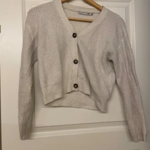 Grå/Beige stickad kofta med knappar - Mysig stickad kofta med v-ringning och stora bruna knappar framtill. Modellen är croppad och har långa ärmar. 