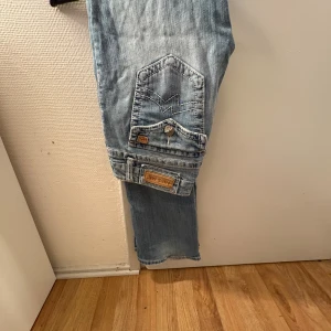 Miss Me ljusblå bootcut jeans, stl 29 - Snygga ljusblå jeans från Miss Me i bootcut-modell med coola detaljer på bakfickan, som broderi och metallknapp. Jeansen har klassisk femficksdesign och är tillverkade i mjukt denim. Perfekt för dig som gillar en avslappnad men trendig stil.