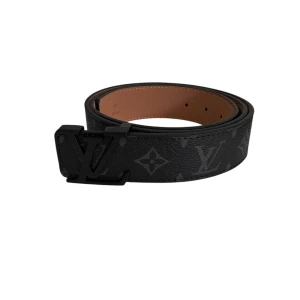 Svart Louis Vuitton bälte med monogram - Snyggt svart bälte från Louis Vuitton med klassiskt monogrammönster och stor LV-logga som spänne. Bältet är gjort i exklusivt skinn och har en mörk, stilren look som passar till jeans eller kostym. Perfekt för dig som gillar lyxiga accessoarer.