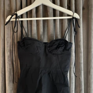 Svart korsett-topp från Zara - Säljer en svart korsett-topp från Zara med tunna axelband som knyts vid axlarna. Toppen har en snygg sweetheart-urringning och lätt rynkade detaljer vid bysten. Materialet är bomull och modellen är croppad med en lätt utsvängd nederdel.