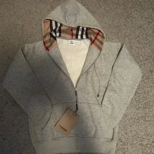 Grå Burberry hoodie med dragkedja - Snygg grå hoodie från Burberry i storlek M med klassiskt rutigt foder i huvan. Tröjan har dragkedja framtill, snörning vid huvan och två fickor. Perfekt för dig som vill ha en stilren och exklusiv look med en touch av Burberrys ikoniska mönster. Säljer pga jag sparar pengar