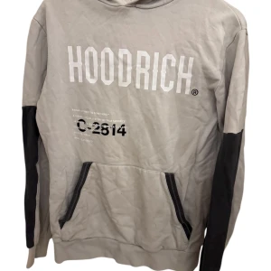Grå hoodie från Hoodrich med tryck - Säljer en skön grå hoodie från Hoodrich med stor vit logga framtill och svart tryck. Tröjan har svarta detaljer på ärmarna, huva och en stor magficka med svarta dragkedjor. Kan gå ner i pris vid snabb affär!!!